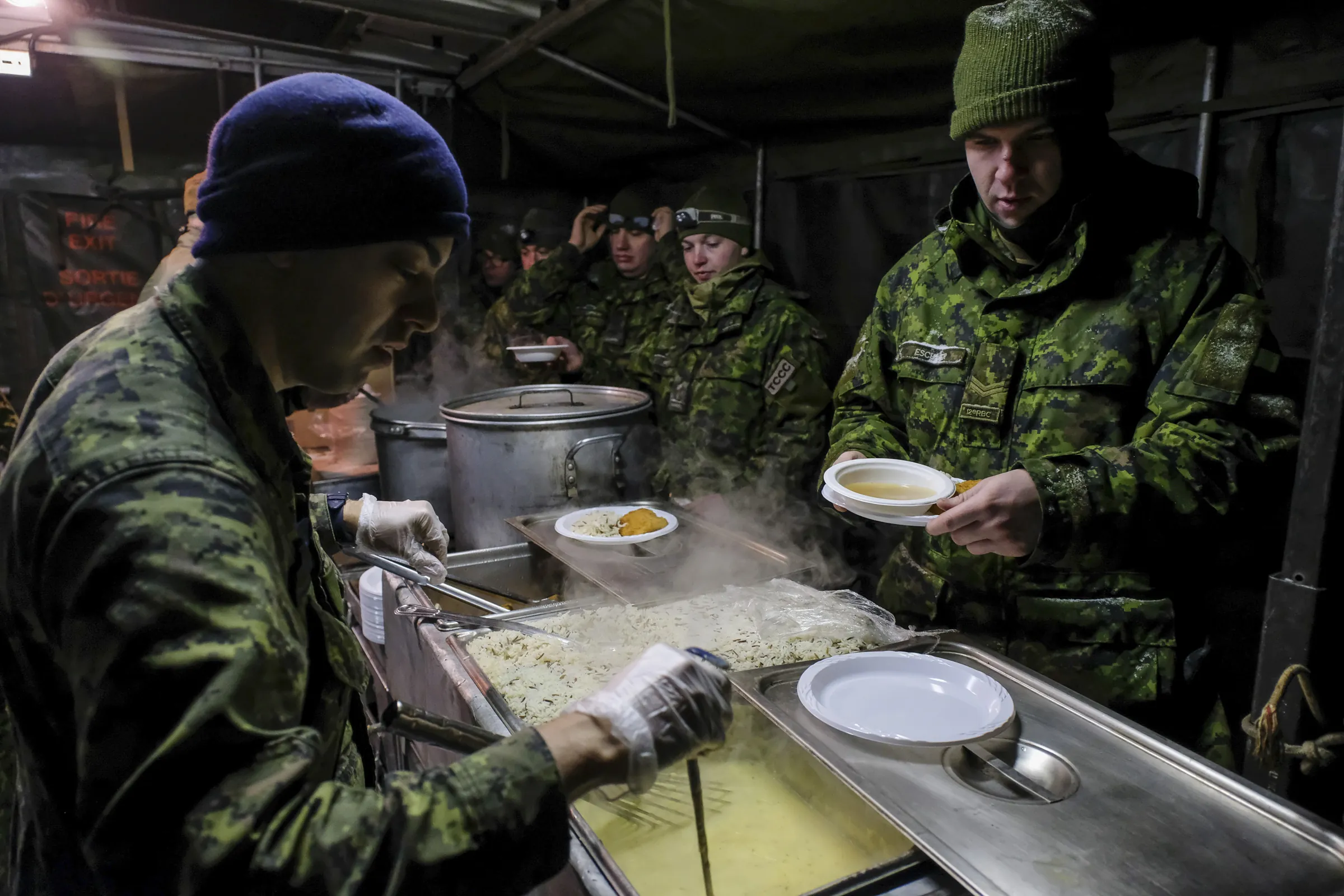 A cook serves hot food to queued soldiers in a field kitchen tent at night