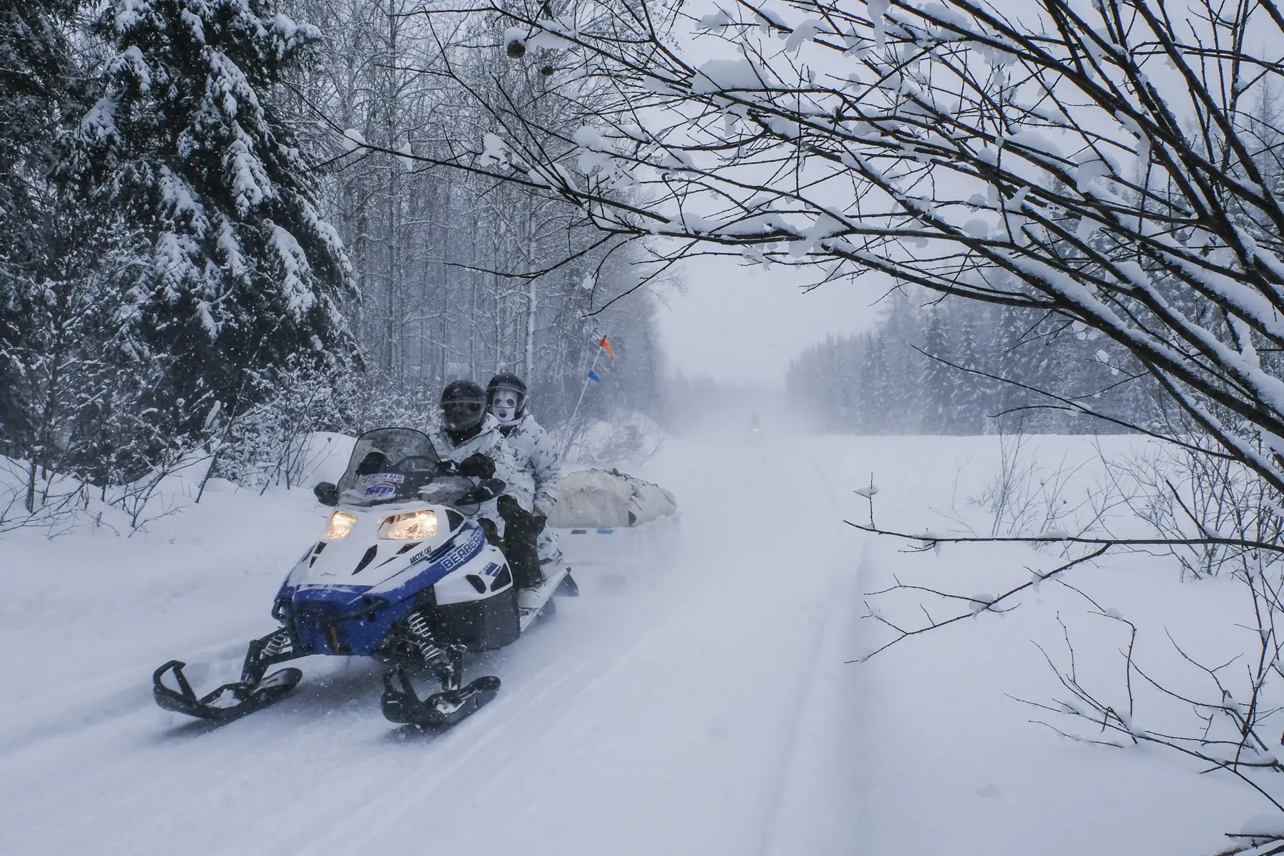 Two soldiers in white camouflage ride a snowmobile with cargo sled through heavy snowfall