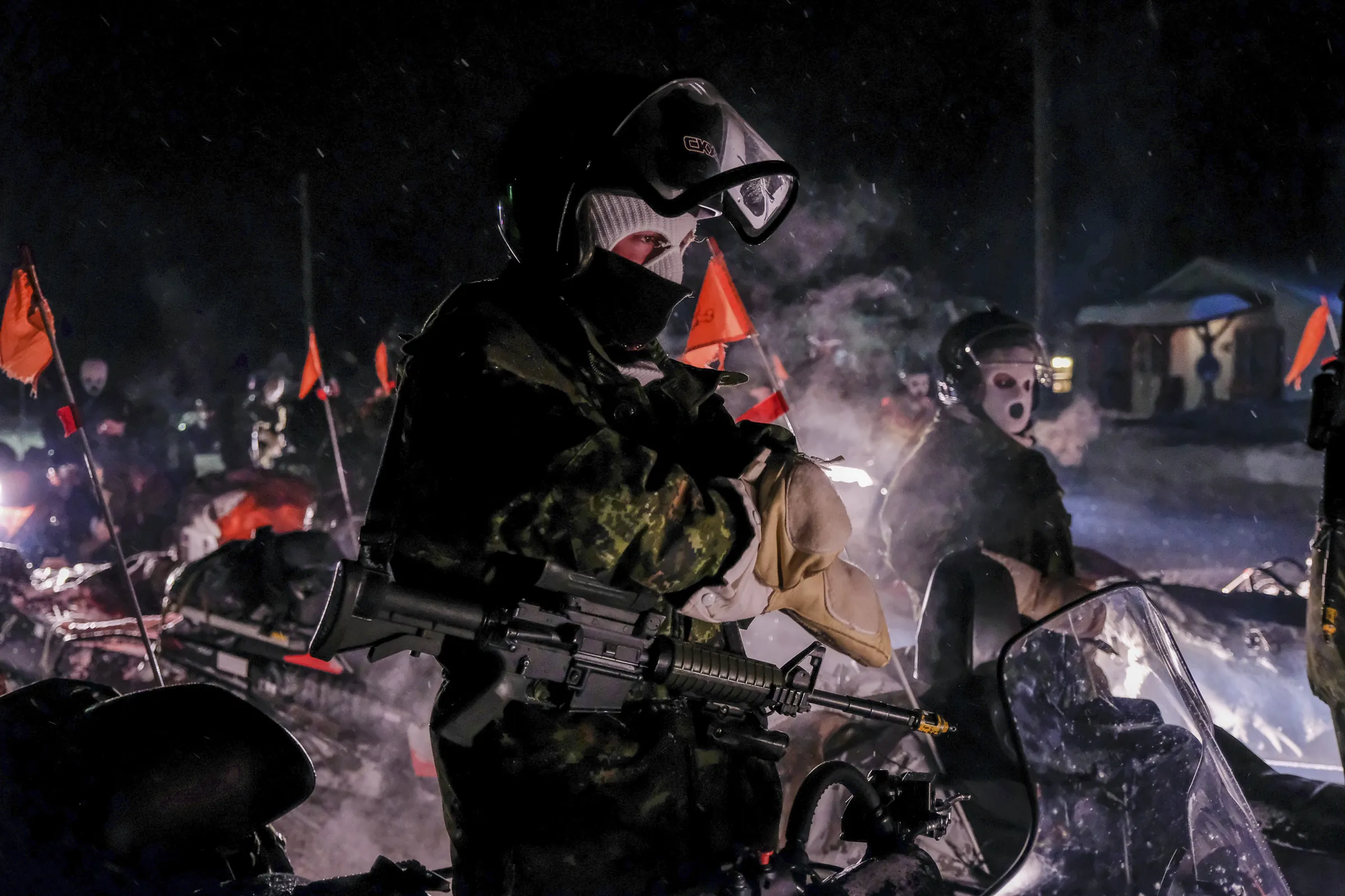 A balaclava-clad soldier with rifle beside snowmobiles at night, faces masked with frost