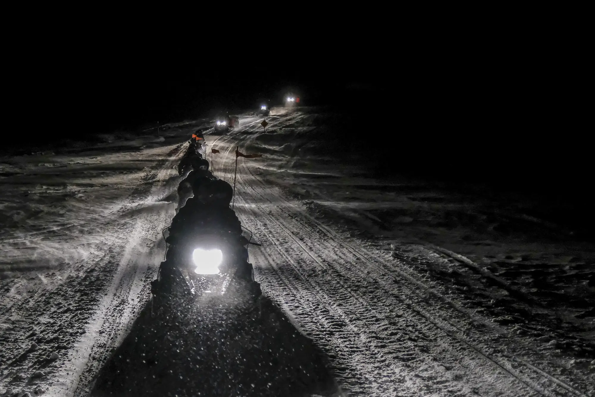 A night snowmobile convoy, headlights strung along a rutted snow trail