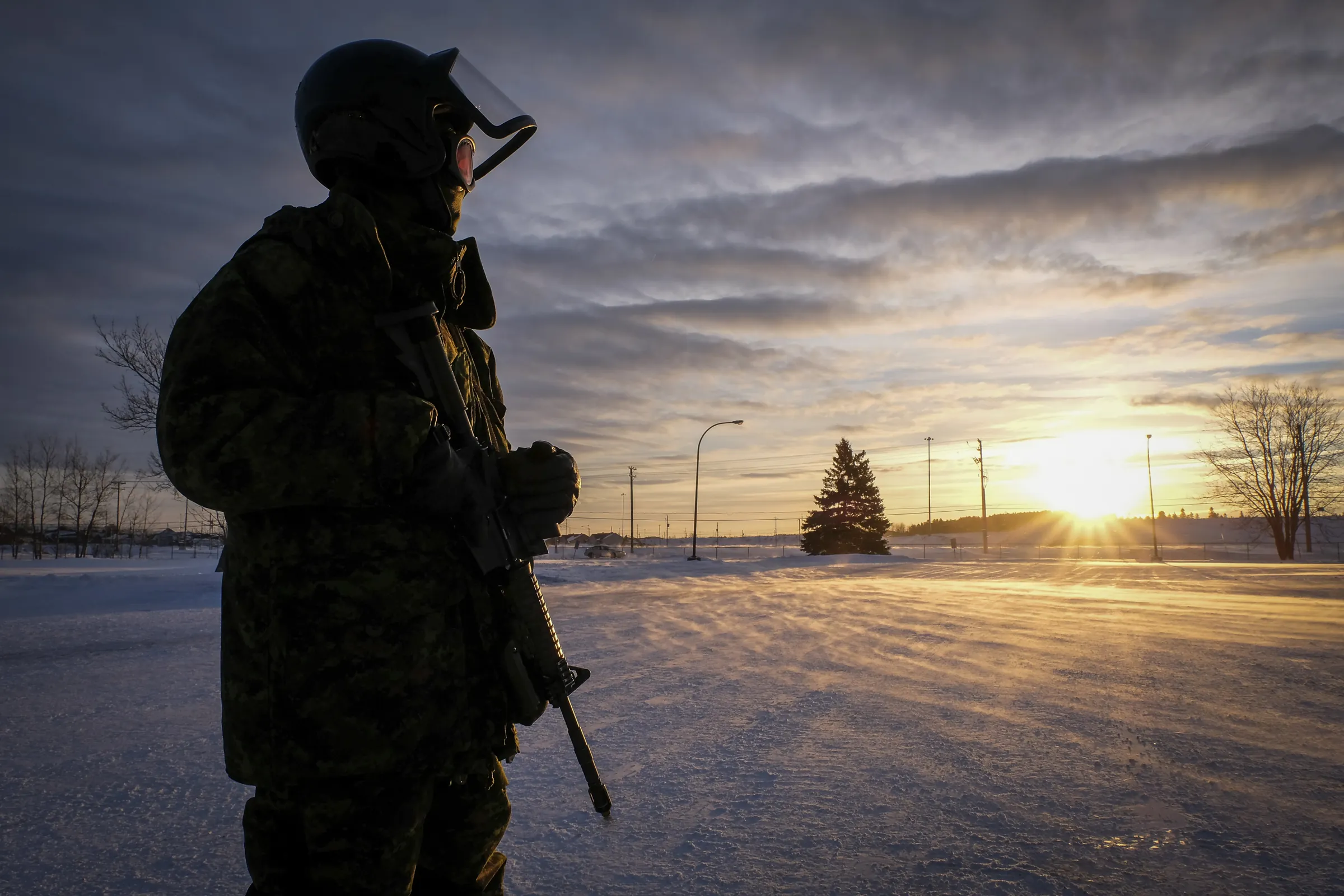 A silhouetted sentry with rifle at golden sunrise over a windswept, icy Canadian camp