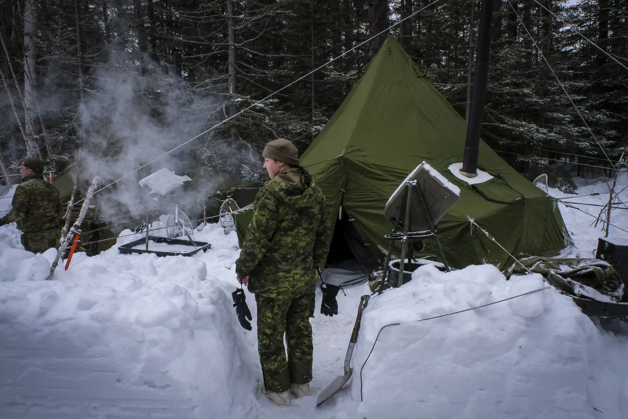 A soldier outside a green arctic tent with smoking stovepipe, snowshoes planted around