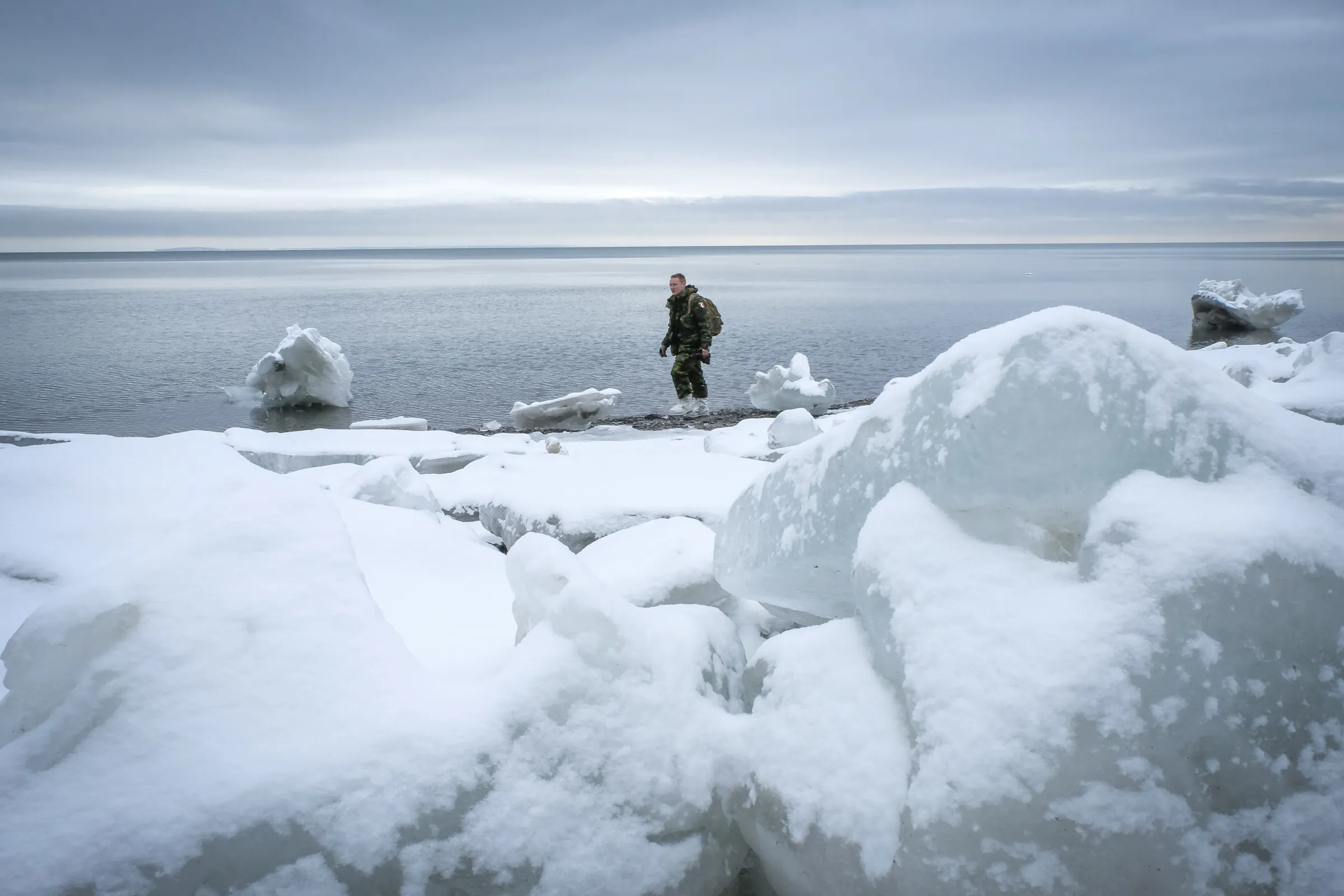 A soldier with a France shoulder patch walks a shoreline between huge snow-covered ice blocks