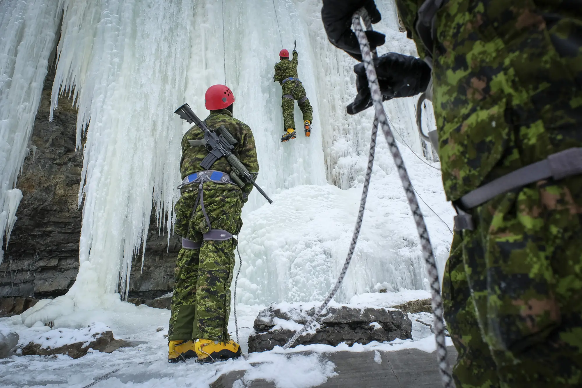 Soldiers ice-climbing a frozen waterfall, a belayer with slung rifle below