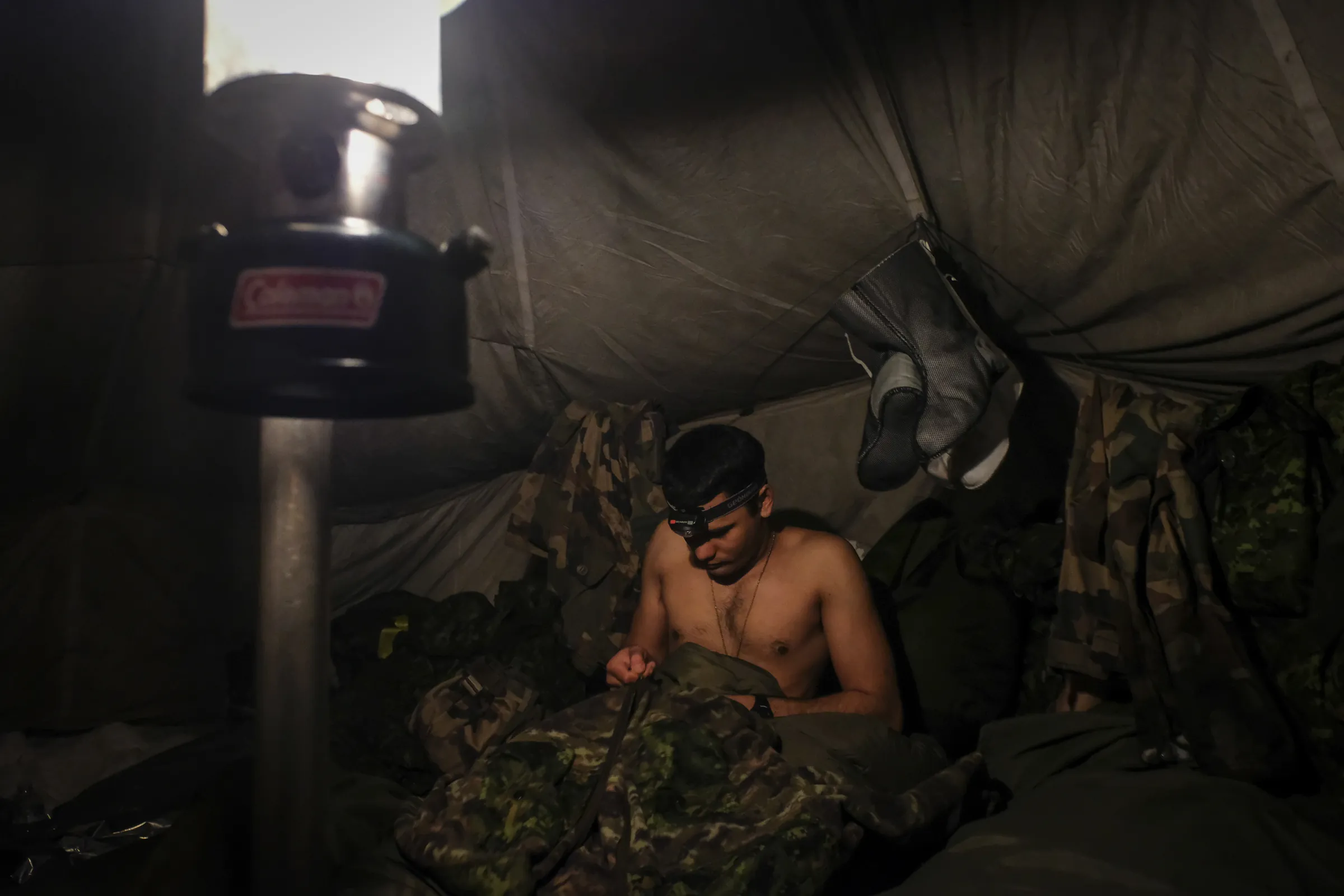 A soldier with headlamp sorts his kit inside a dim tent beside a lantern