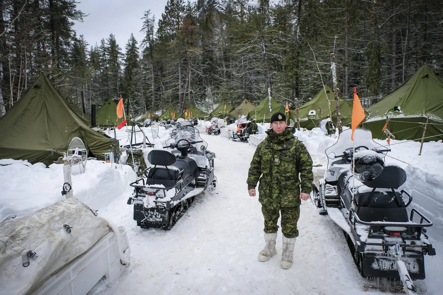 A Canadian officer on a snow path between snowmobiles and rows of green tents