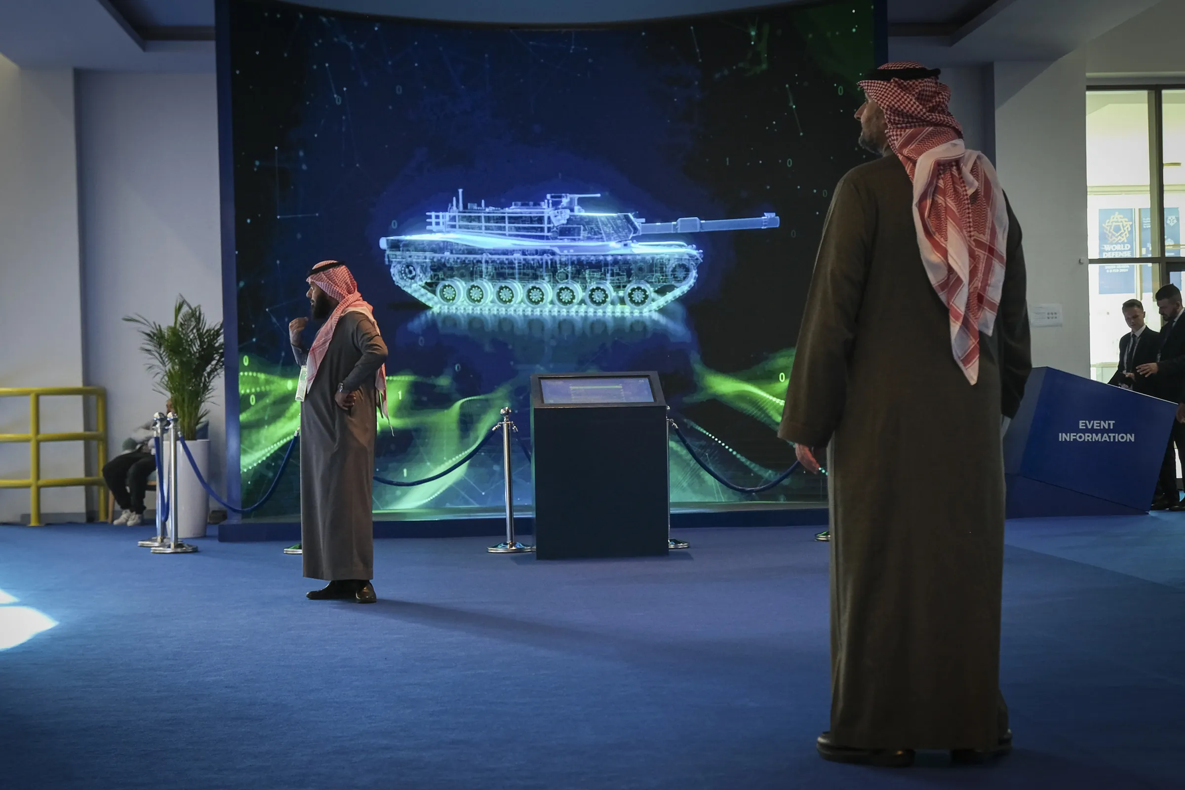 Two visitors in Saudi dress watch a glowing holographic tank on a giant LED wall in a defence trade show lobby