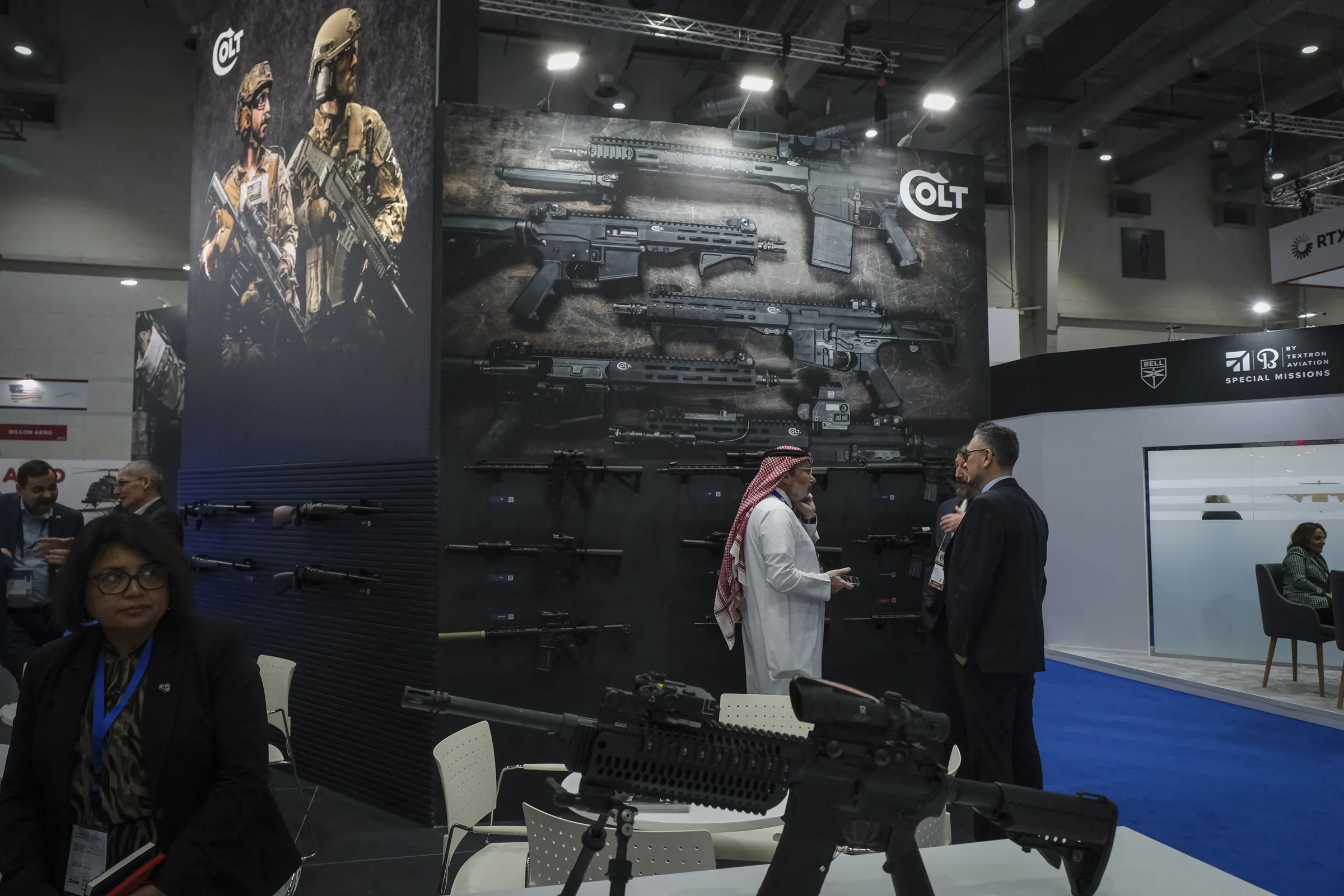 A visitor in a white thobe talks with Western executives at a stand of wall-mounted rifles
