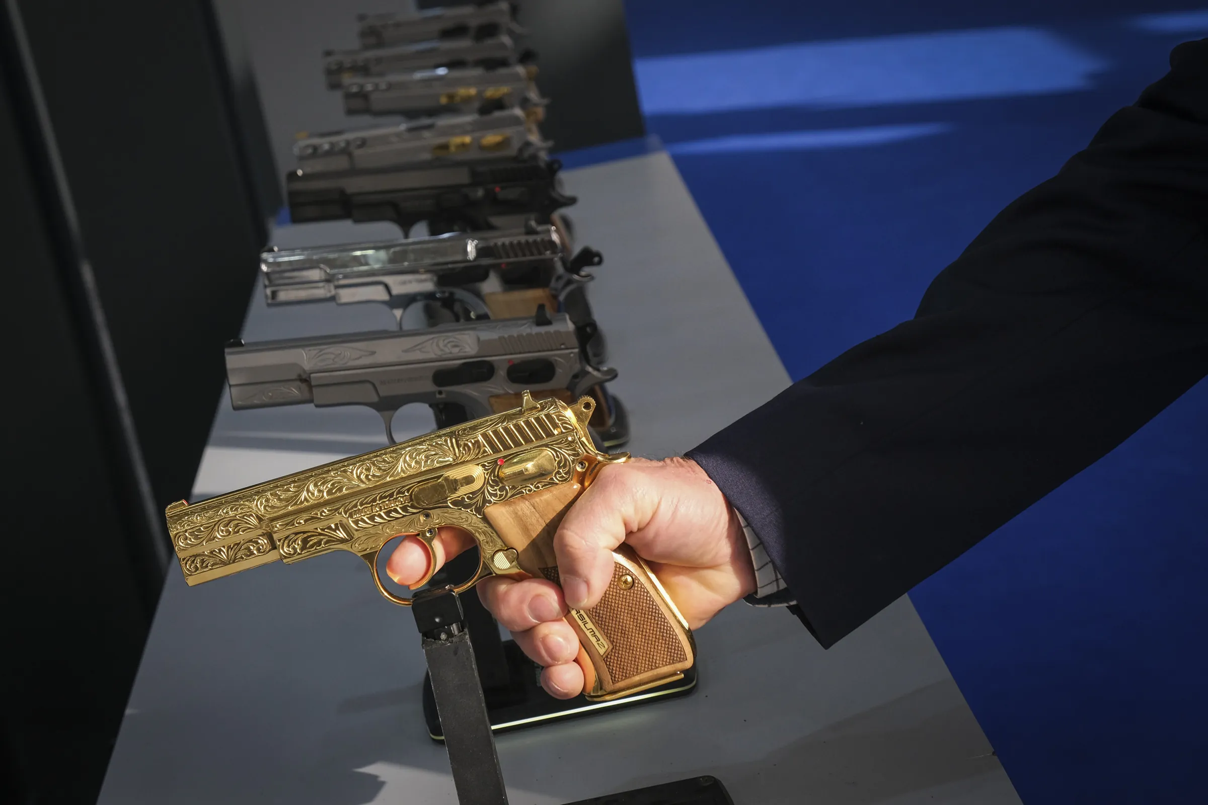 A suited buyer grips an ornately engraved gold-plated pistol on a display rail