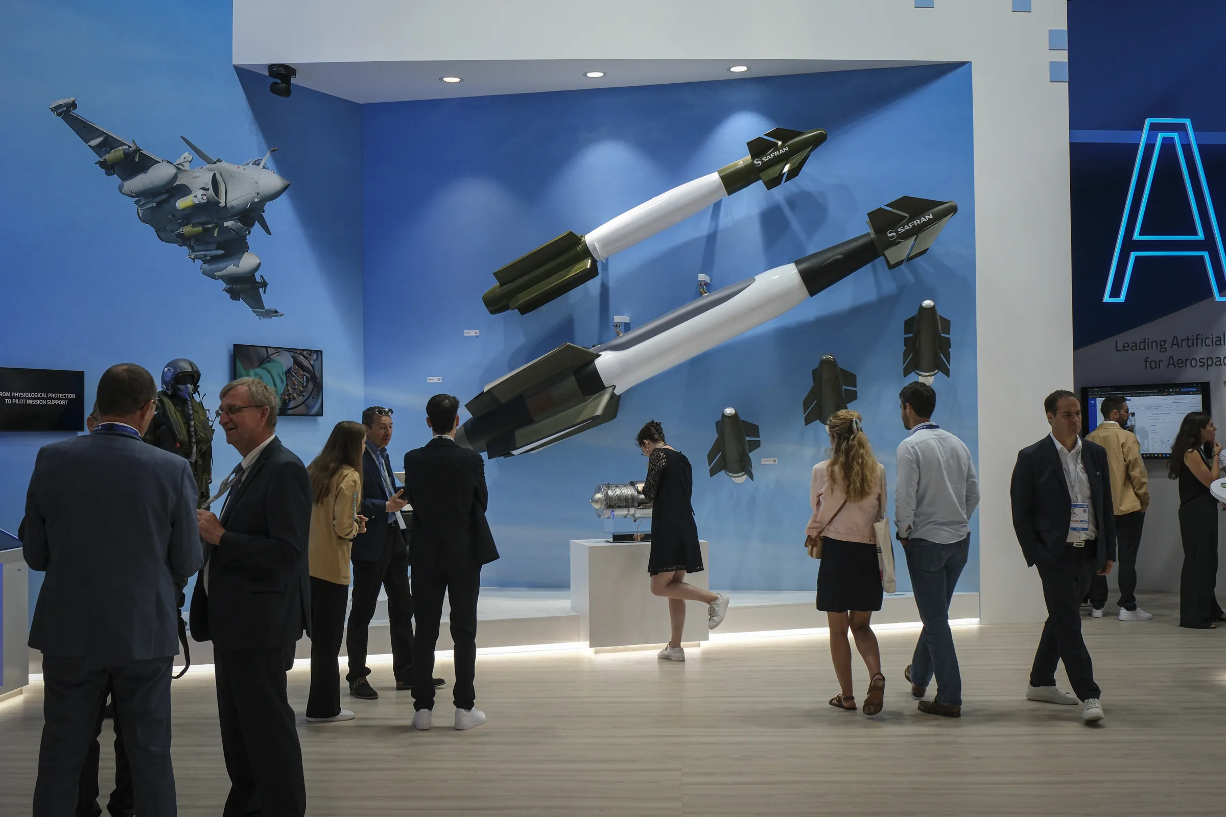 Two large guided bombs jut from a sky-blue wall with a fighter jet mural as visitors mingle