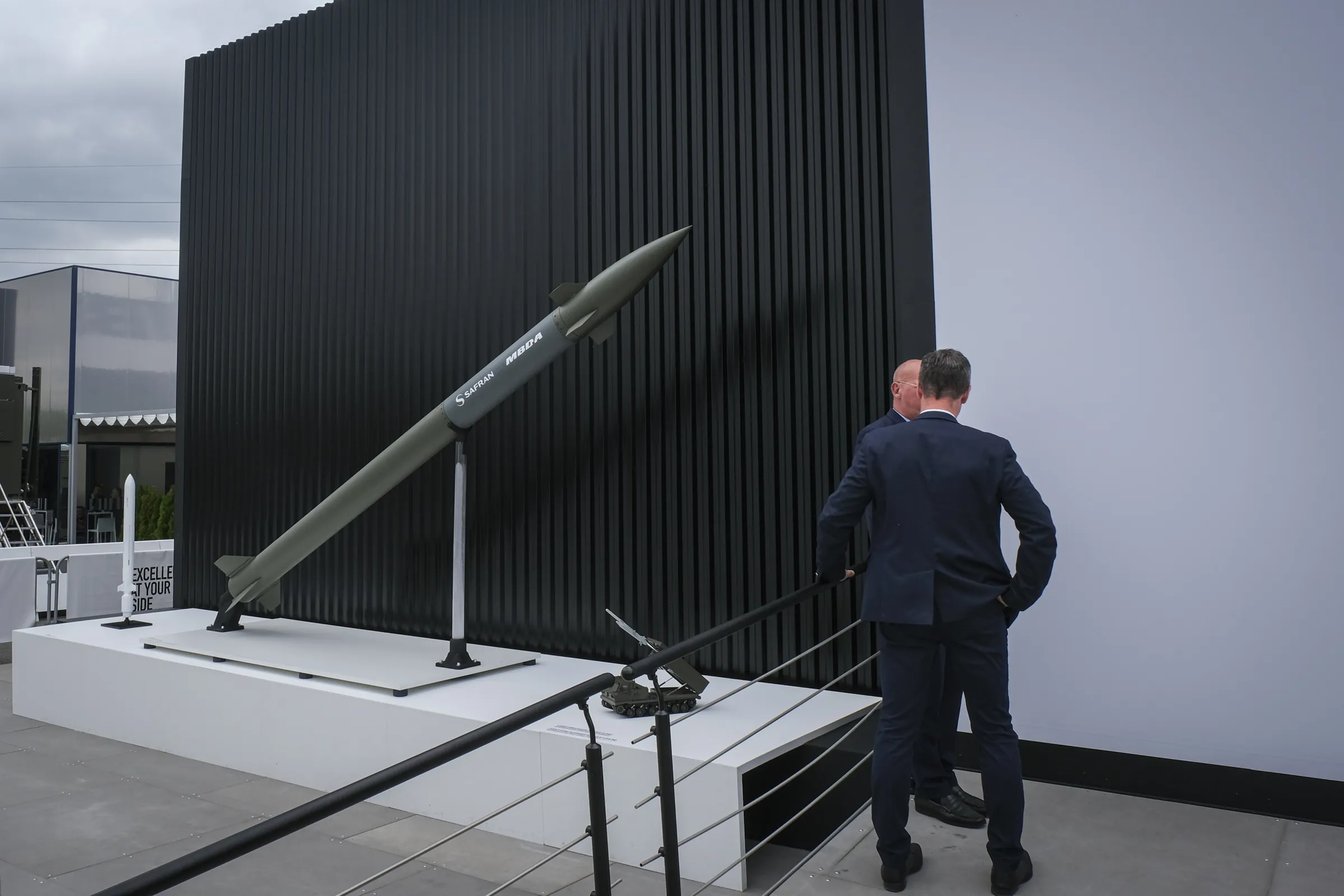 Two executives contemplate an upright missile against a black slatted wall under a grey sky