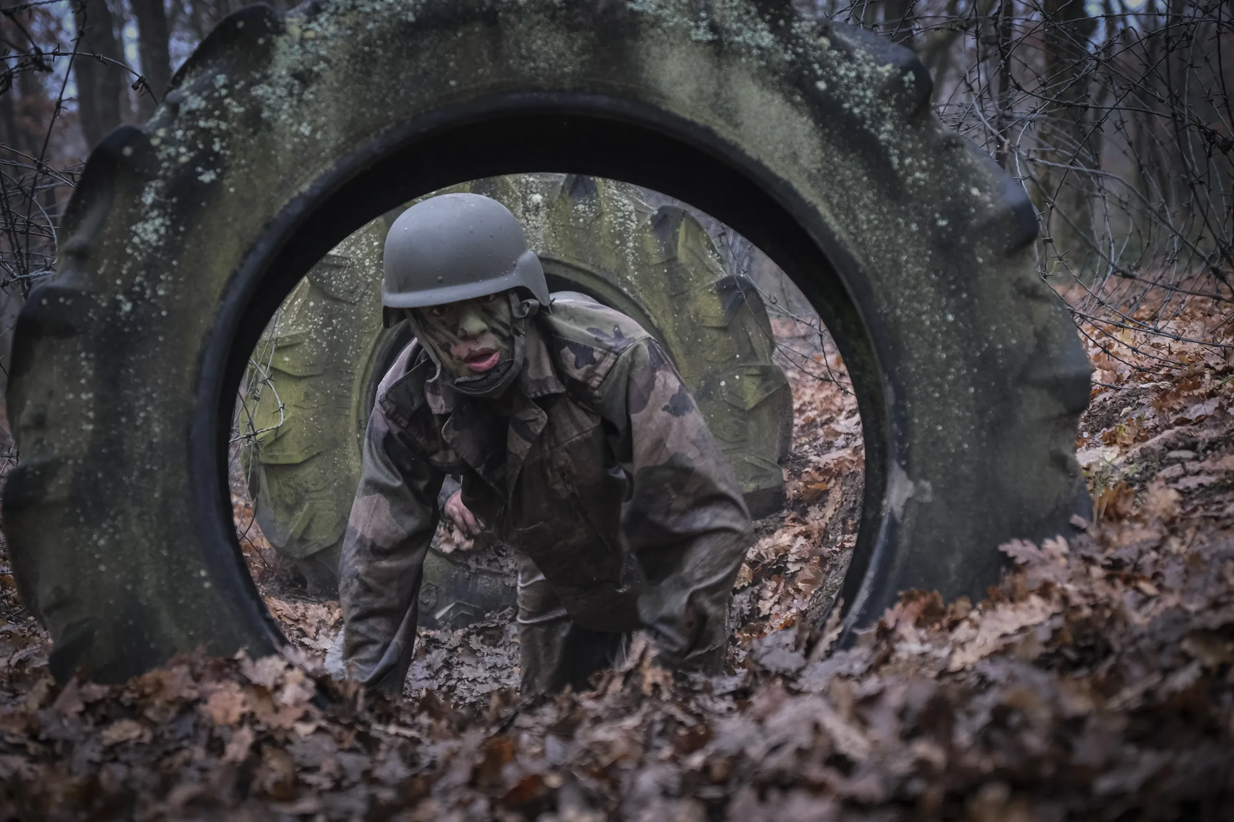 A recruit with camouflage face paint crawls through a giant tyre obstacle under barbed wire