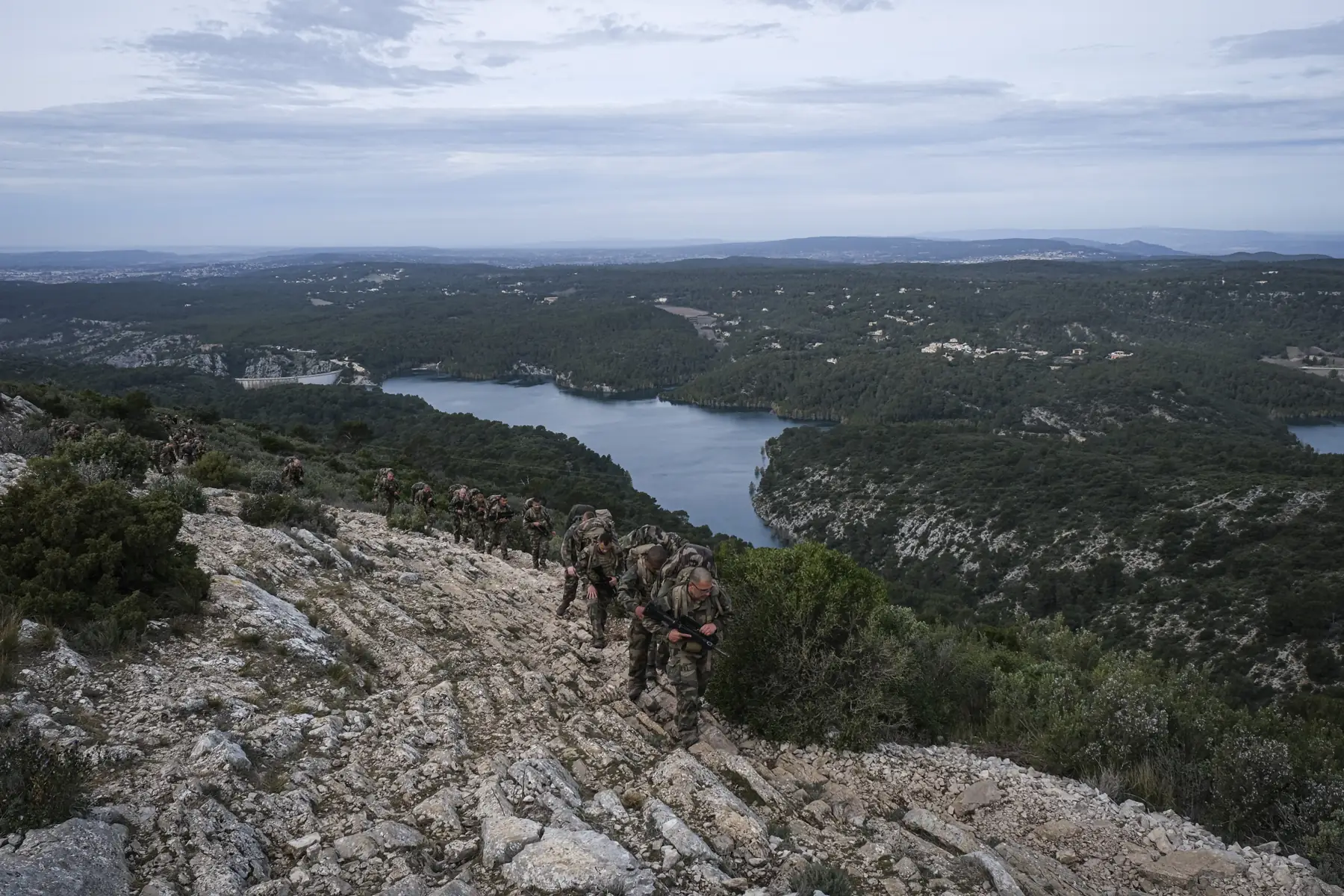 A long file of loaded Foreign Legion recruits climbs a rocky ridge above a lake