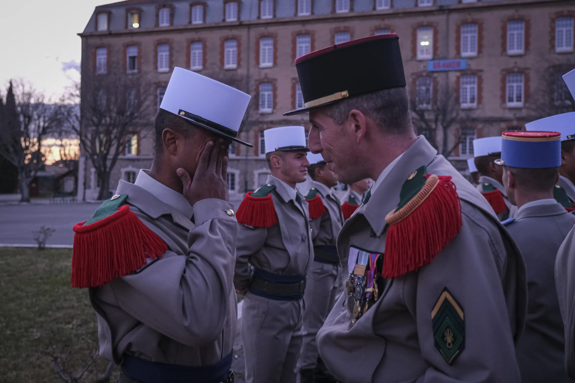A new legionnaire in white képi wipes away tears facing a decorated officer at twilight