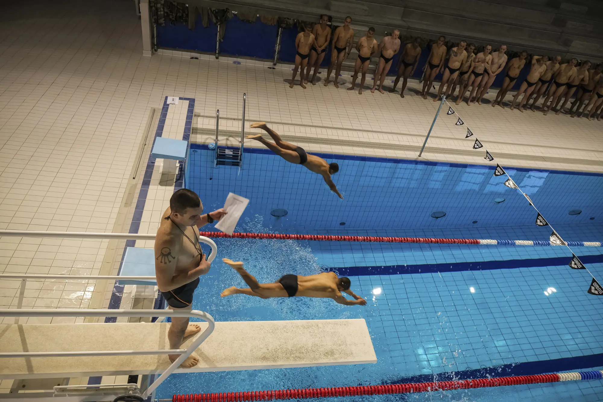 Foreign Legion recruits dive into an indoor pool during a swim test, a long line waiting