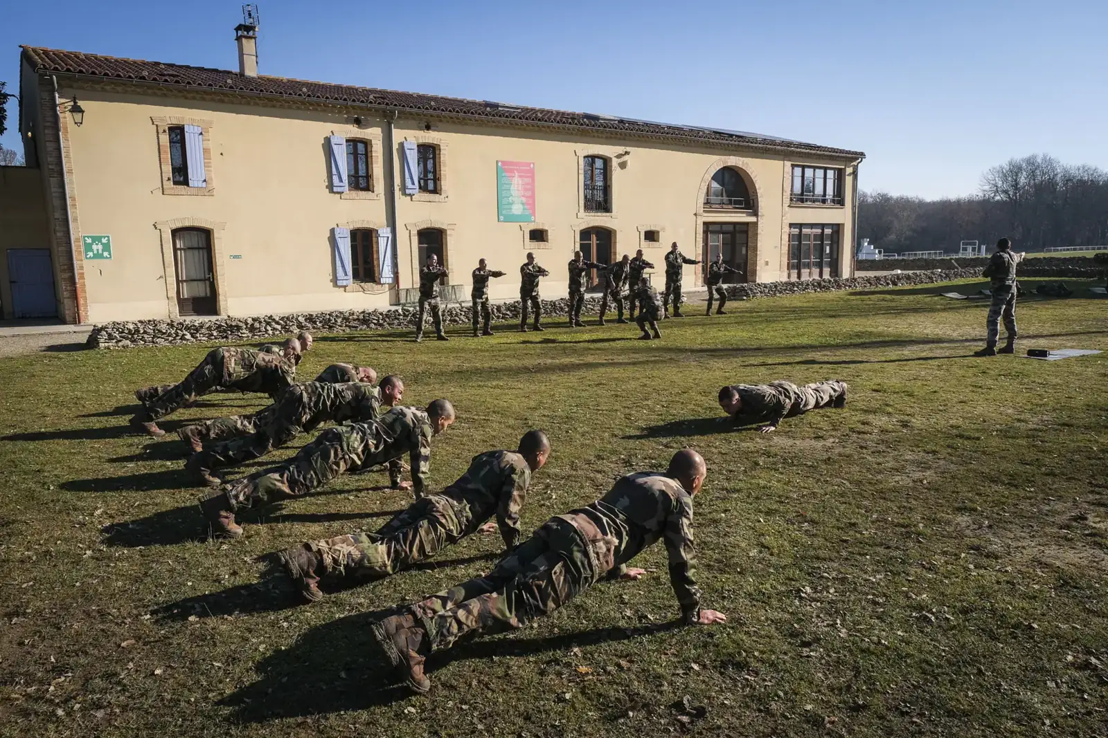 Rows of Foreign Legion recruits do push-ups on the grass before an ochre Provençal farmhouse