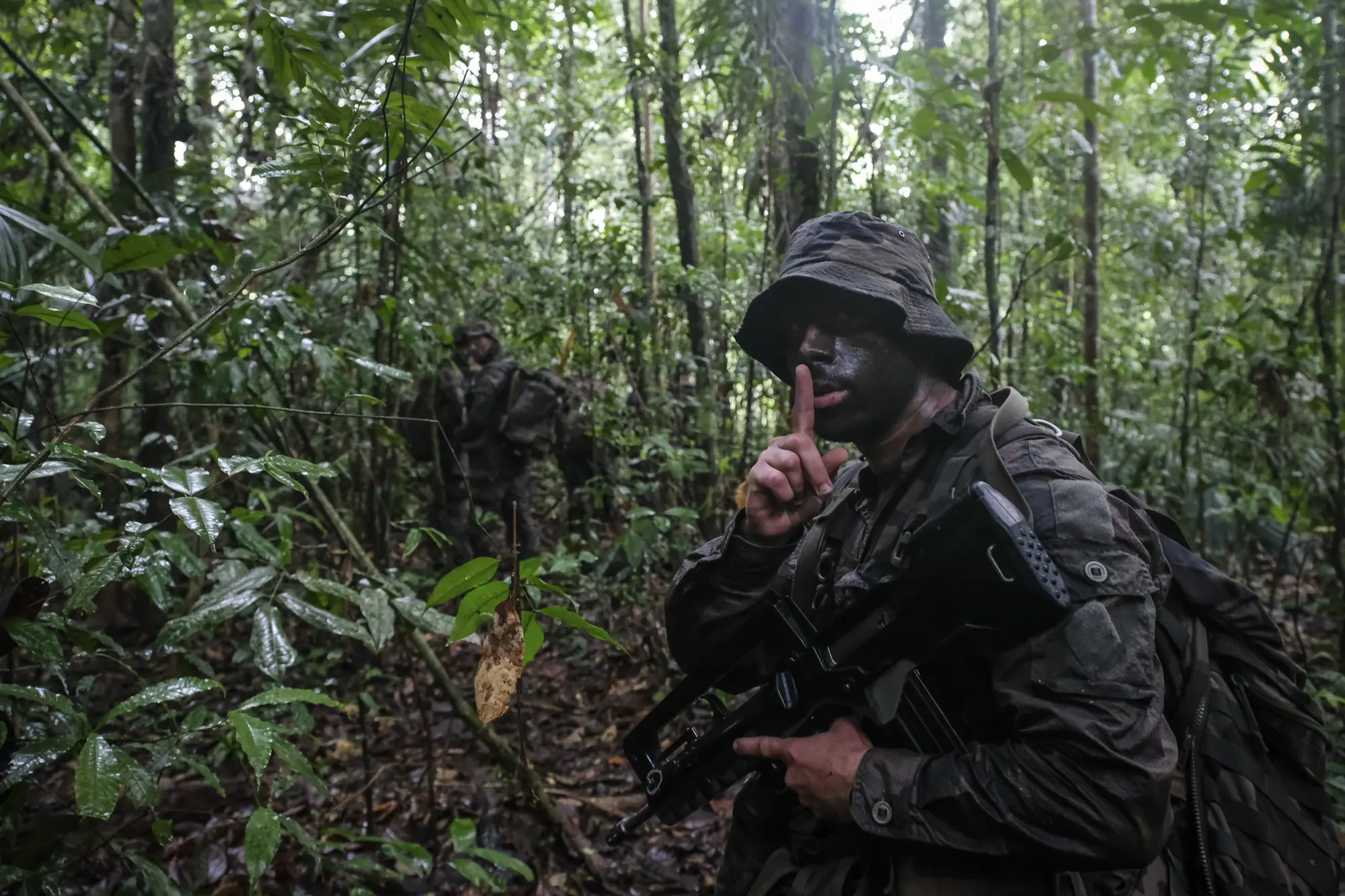 A camouflaged French soldier raises a finger for silence on a dense rainforest trail in French Guiana