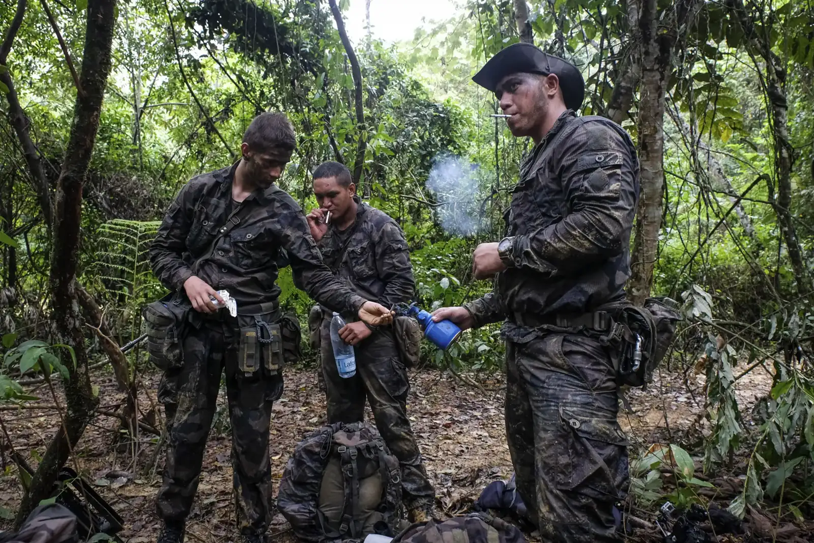 Three mud-soaked French soldiers pause in the jungle to smoke and share water