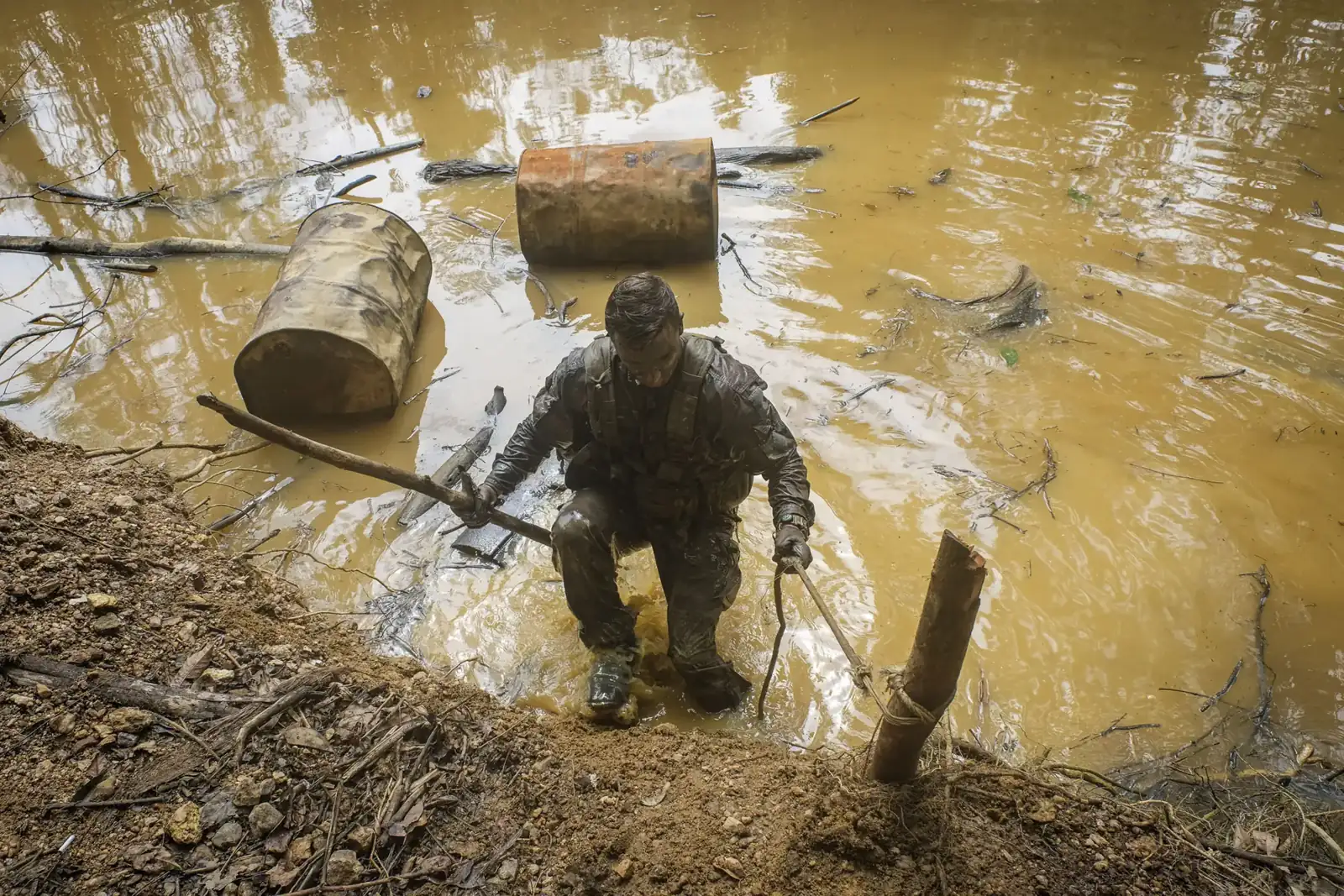 A French soldier climbs out of a yellow, polluted illegal gold-mining pond in French Guiana, rusted fuel barrels floating behind him