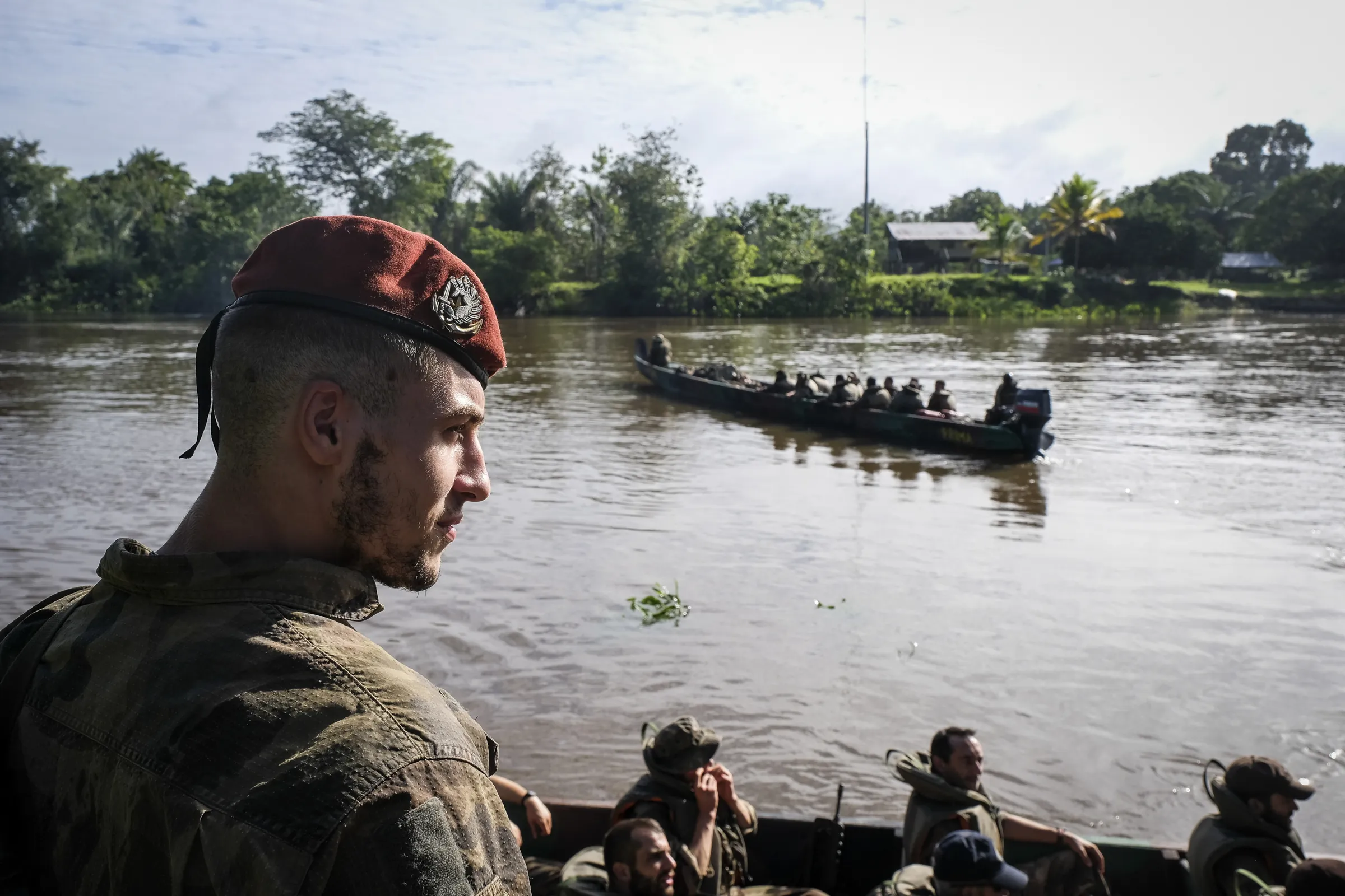 A French Army paratrooper in a red beret watches a troop-filled pirogue cross the river