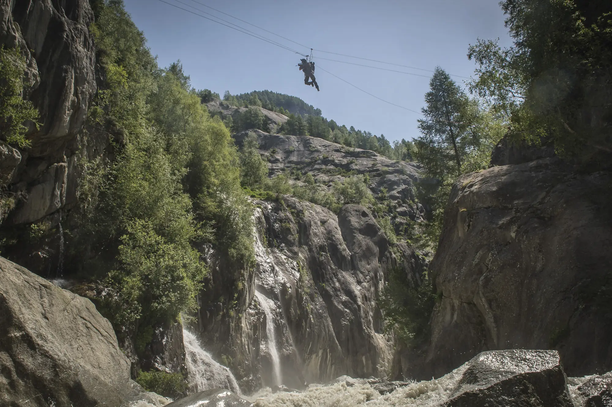 An armed soldier crosses a tyrolean line high above a gorge with waterfalls