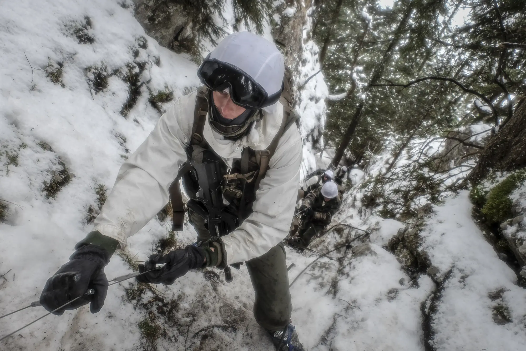 A soldier in white camouflage climbs a steep snowy gully on a fixed cable