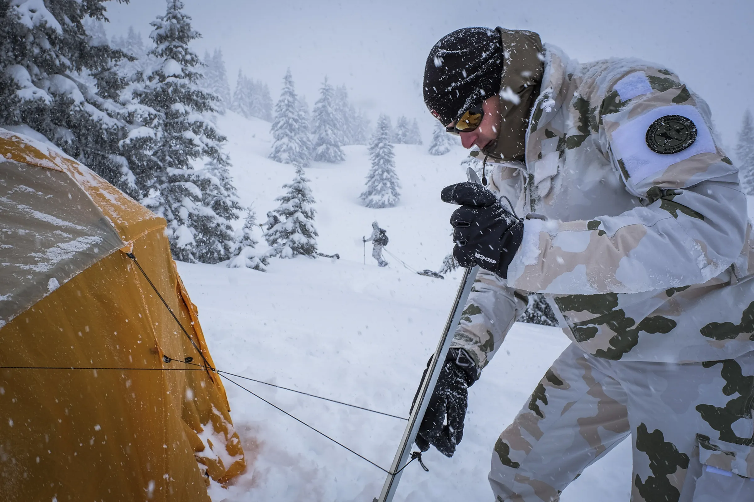 A soldier anchors a tent with a ski during heavy snowfall, another hauling a sled behind