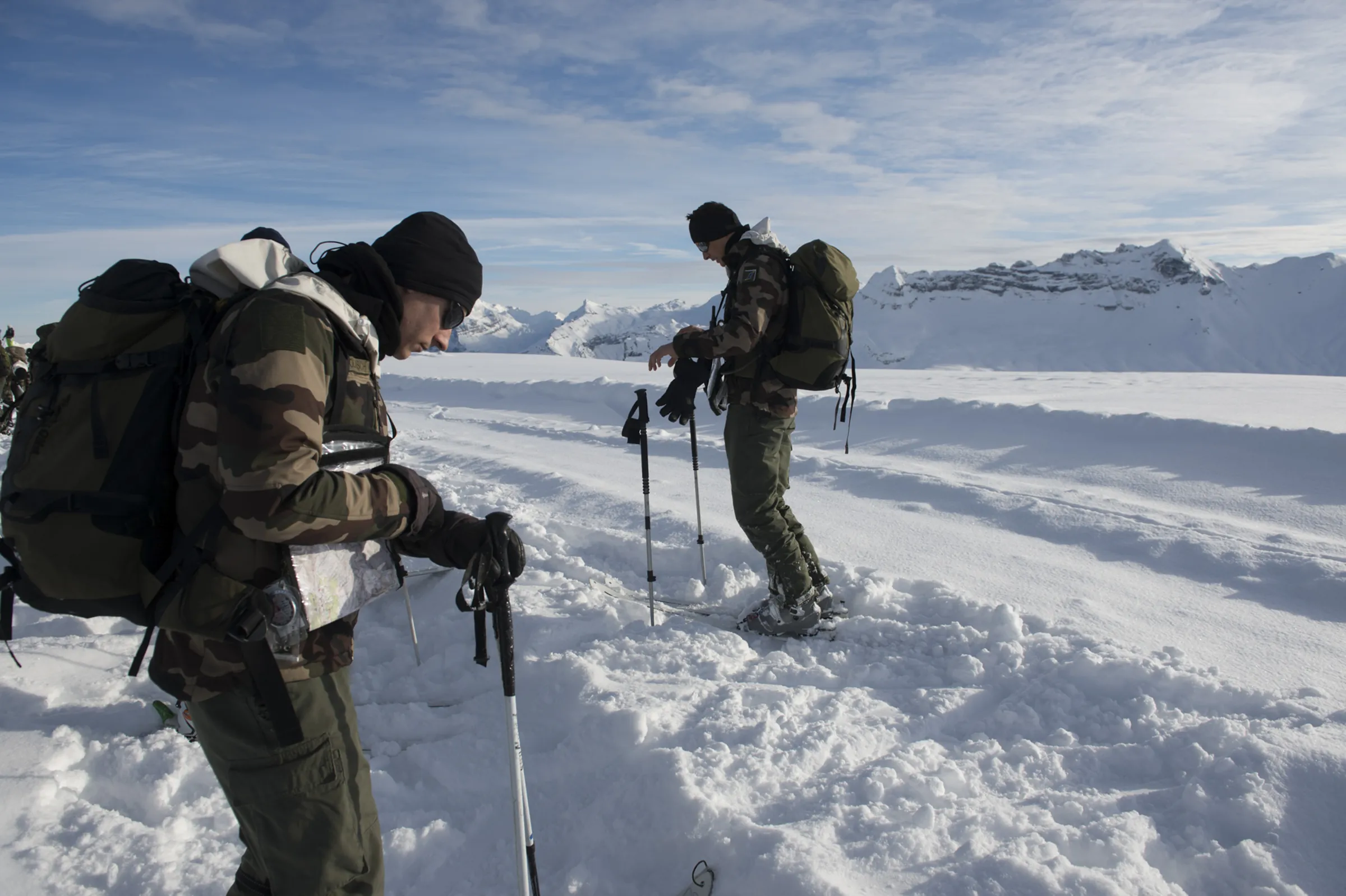 Two French mountain soldiers set off on a long mountain run over a sunlit snowy col, peaks behind