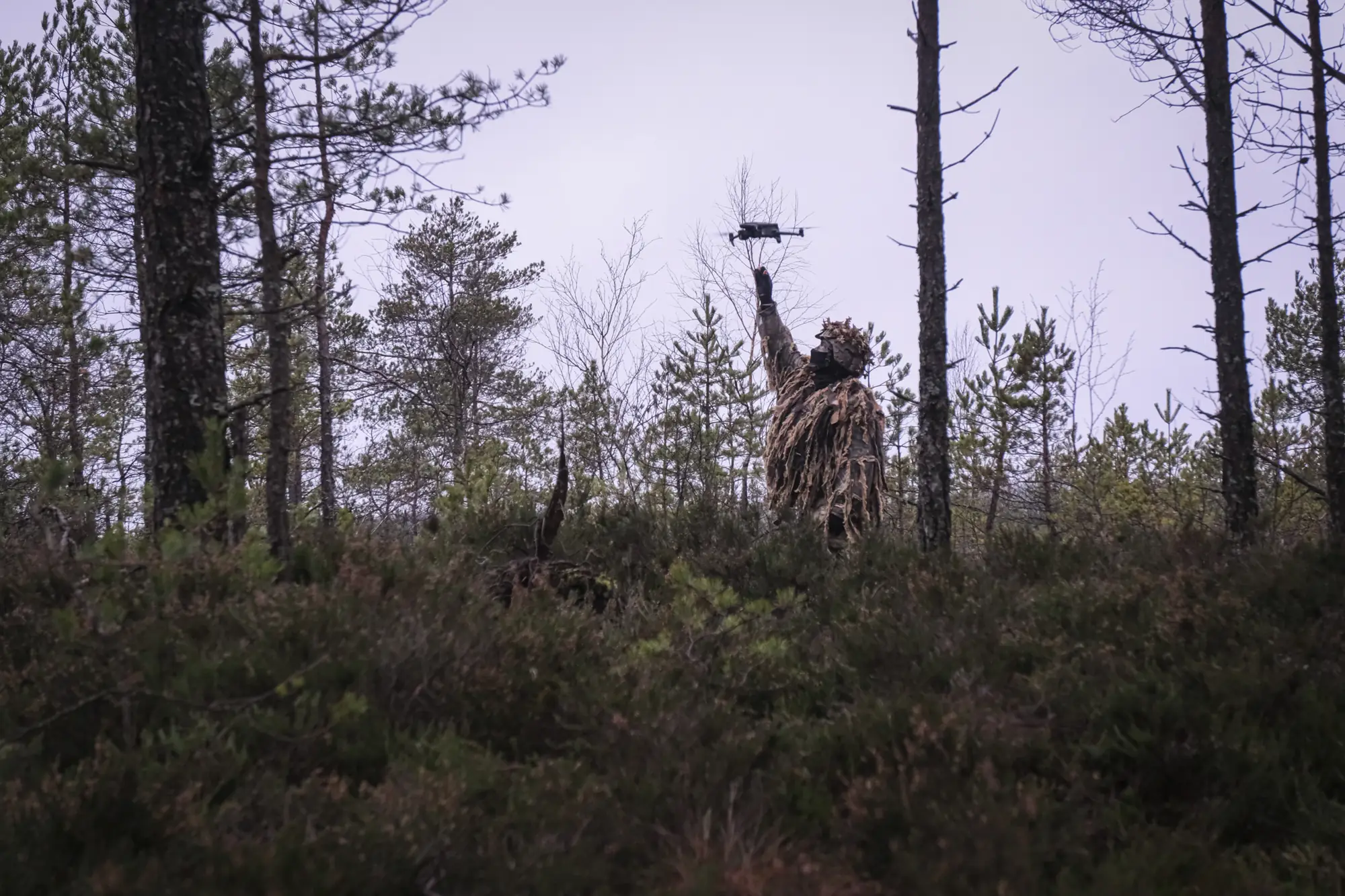 A ghillie-suited sniper raises a hand toward a small drone in a sparse pine bog