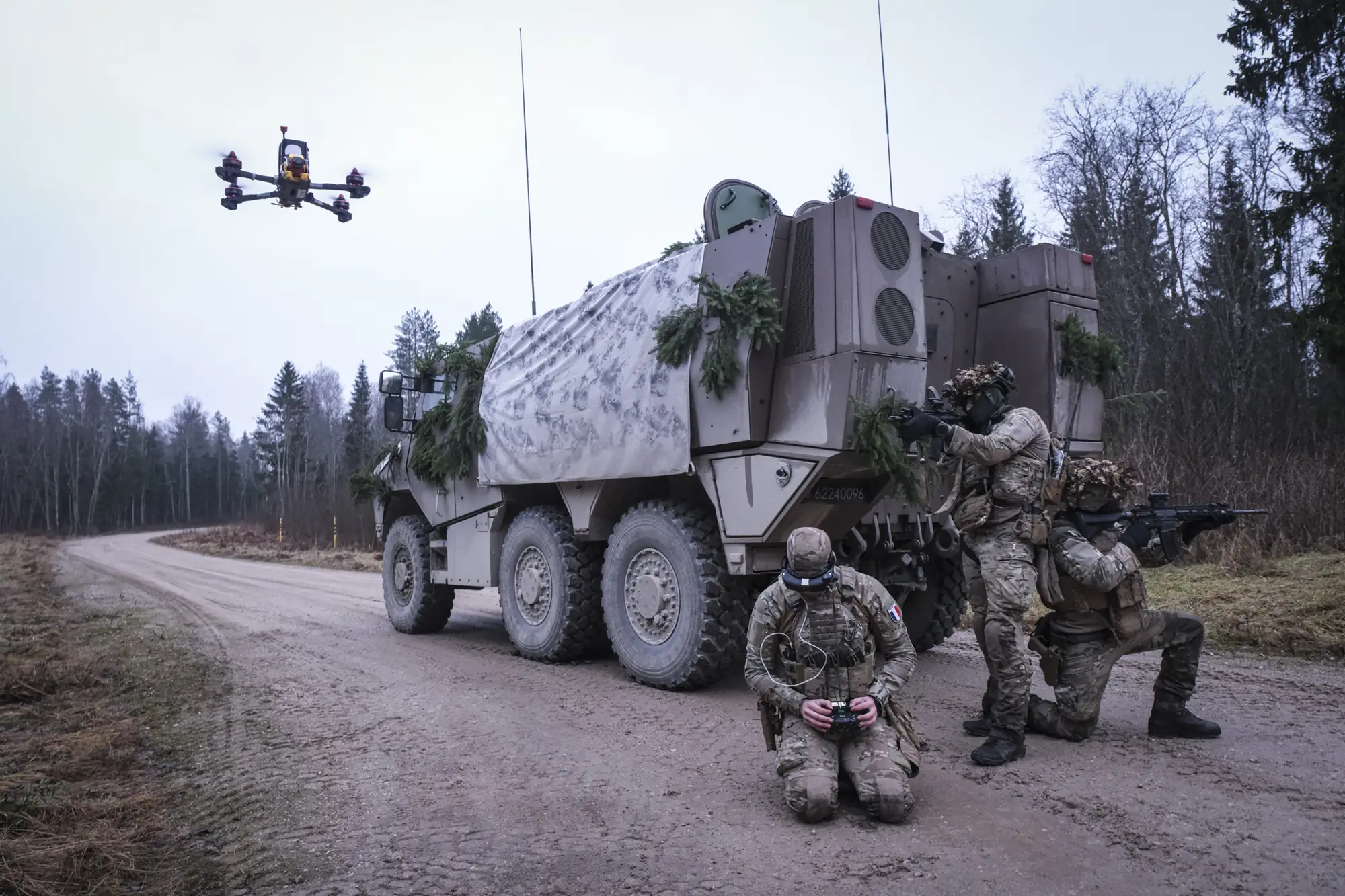 A drone hovers over a French Griffon armoured vehicle covered in snow camouflage and spruce boughs on a misty Estonian road