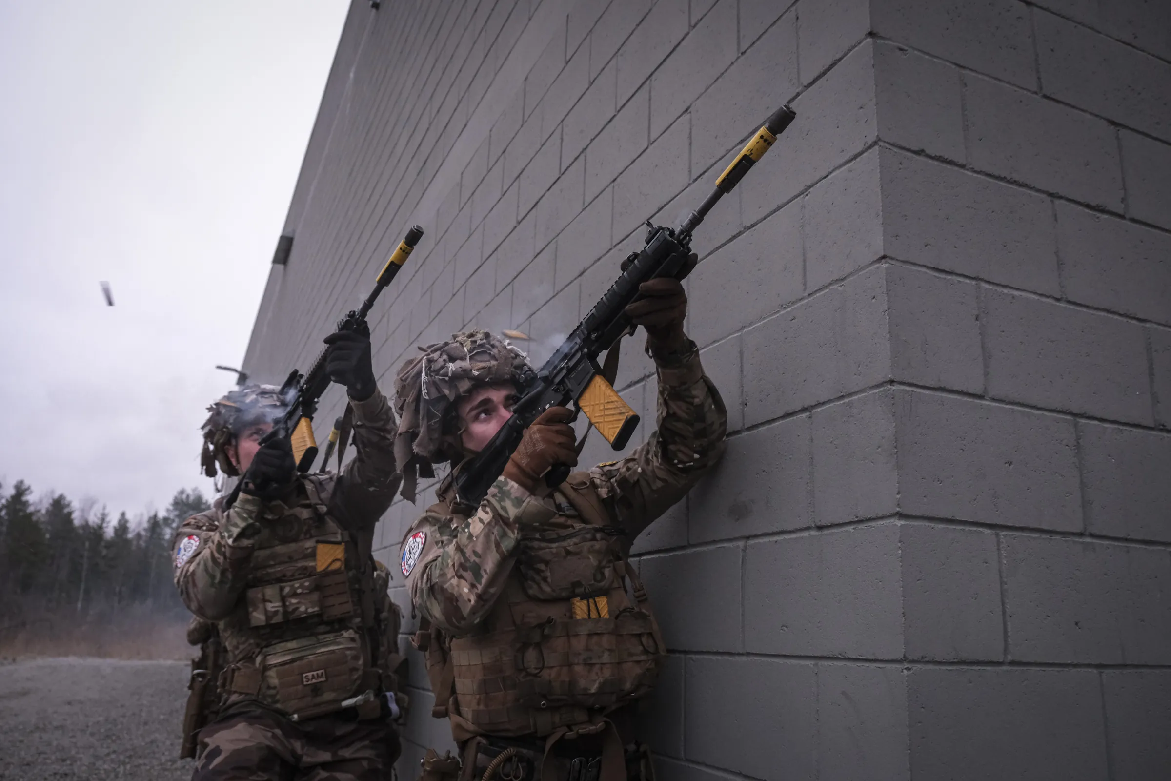Two French soldiers fire rifles along a grey blockhouse corner, ejected casings in the air