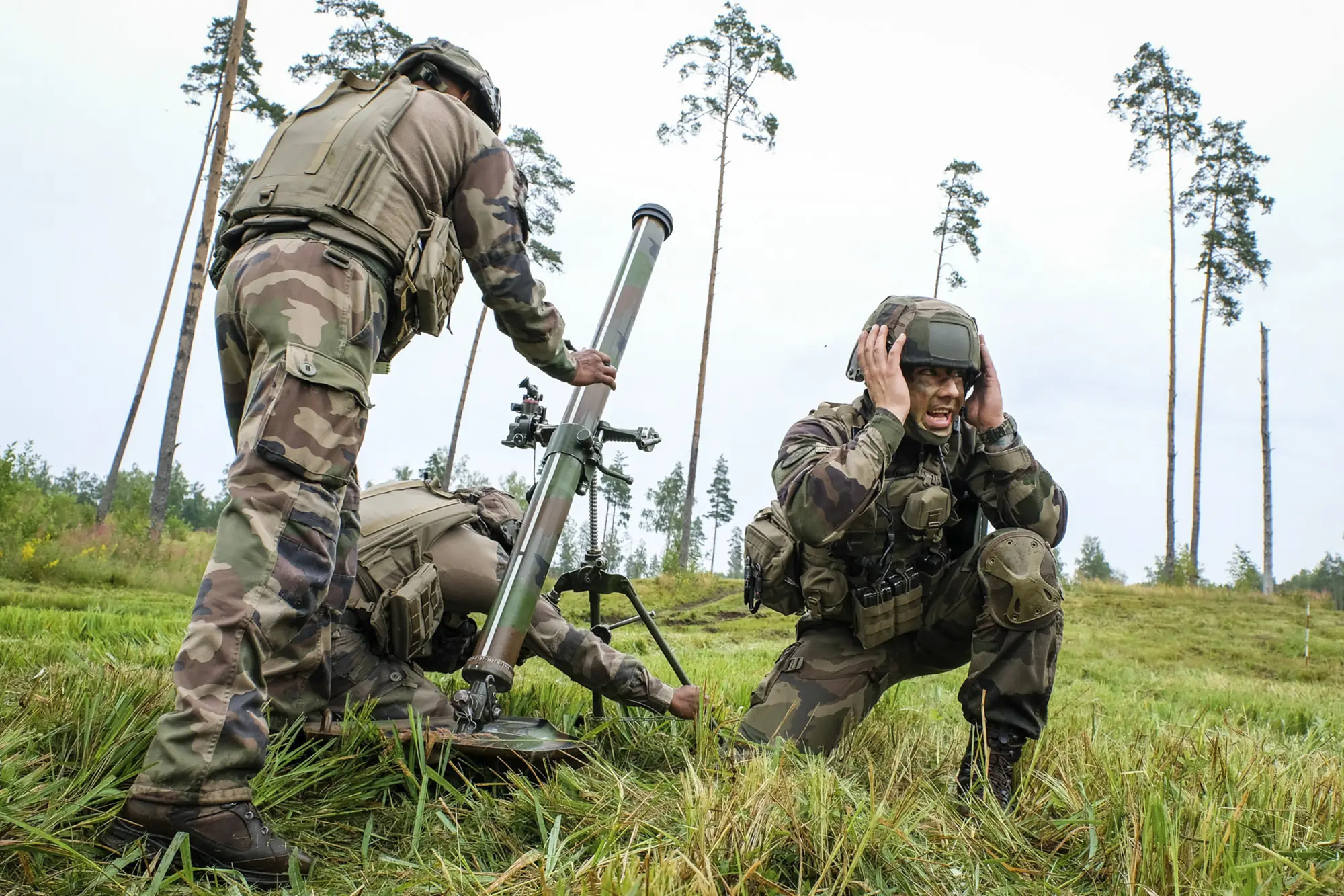 A French mortar team in a green clearing, one soldier covering his ears and shouting