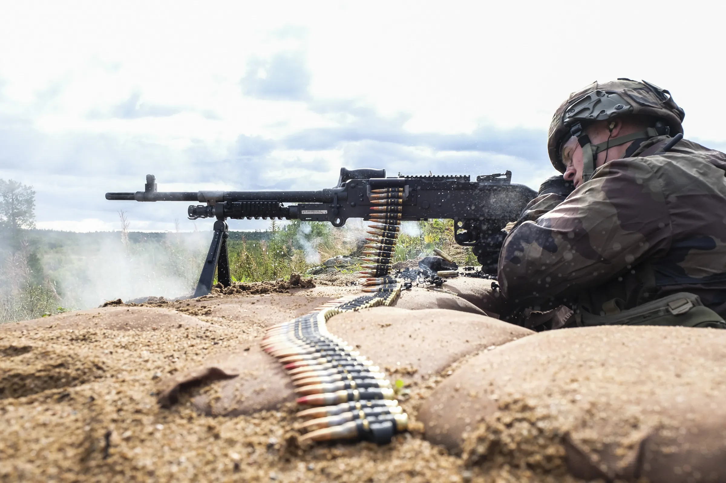 A machine gunner fires a belt-fed machine gun from a sandbagged position