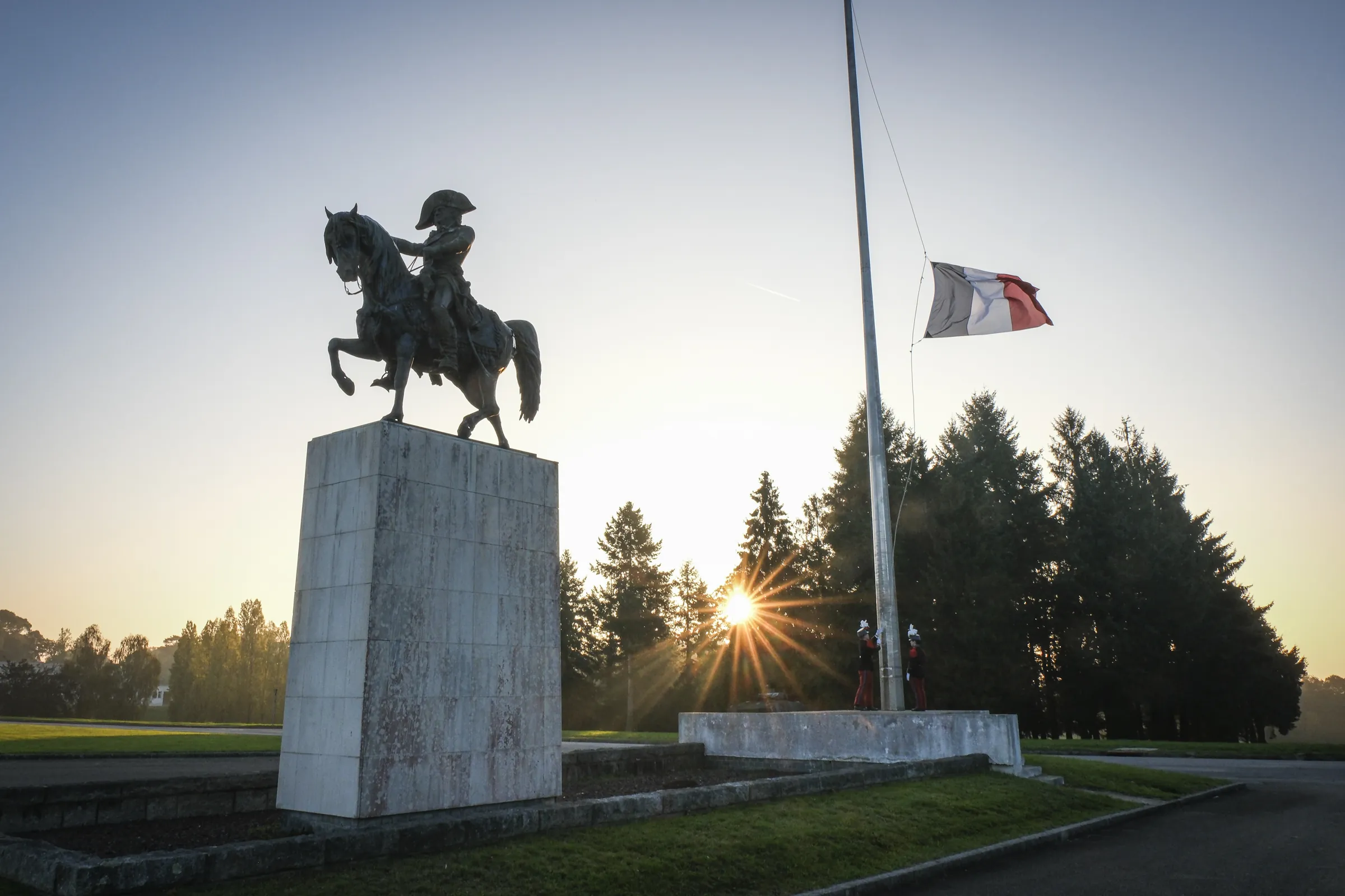 At dawn, two Saint-Cyr cadets in Grand Uniforme raise the French flag beside an equestrian statue, sunburst through the pines