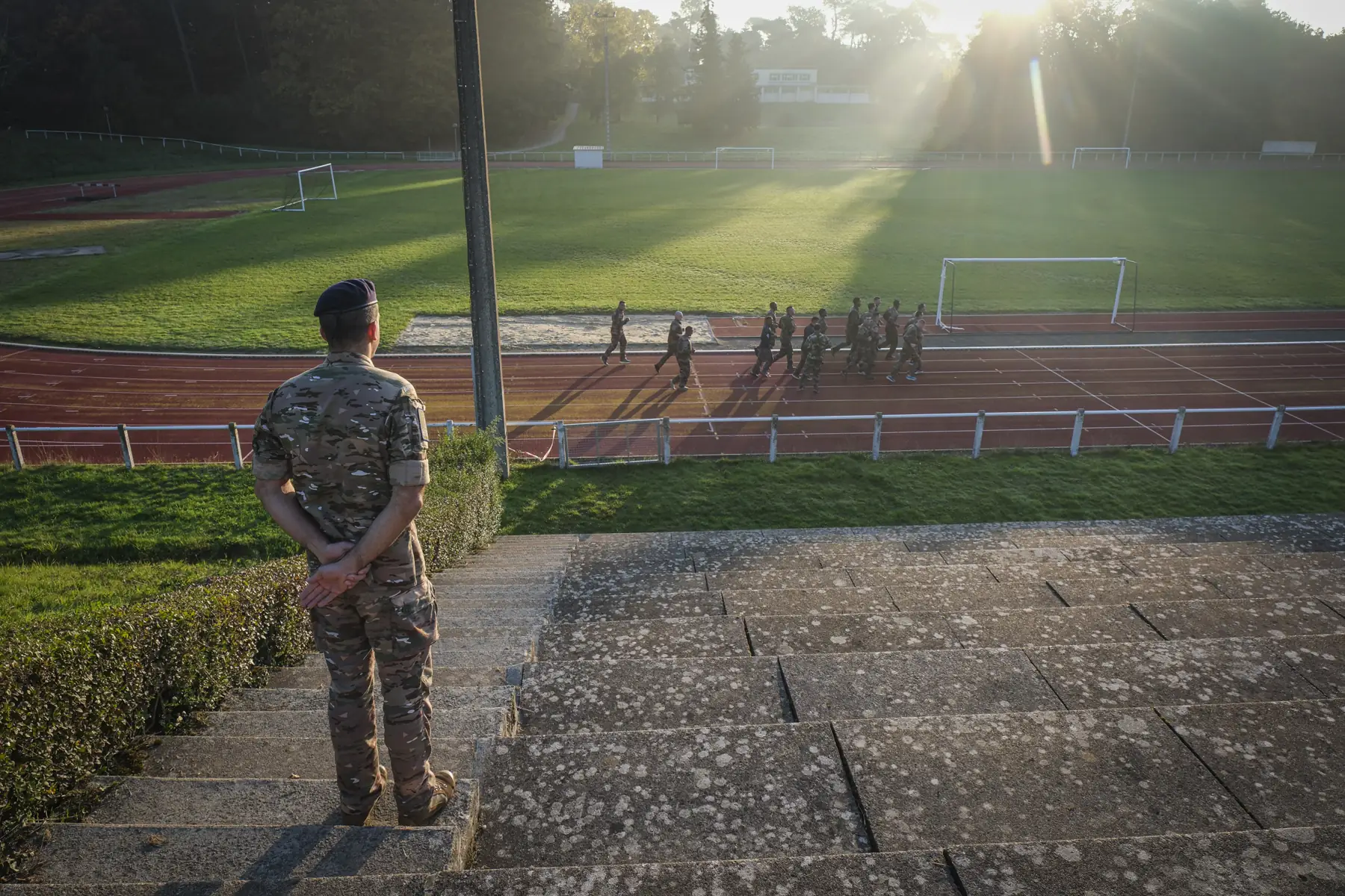 A camouflaged instructor watches from stadium steps as a cadet platoon runs the track below