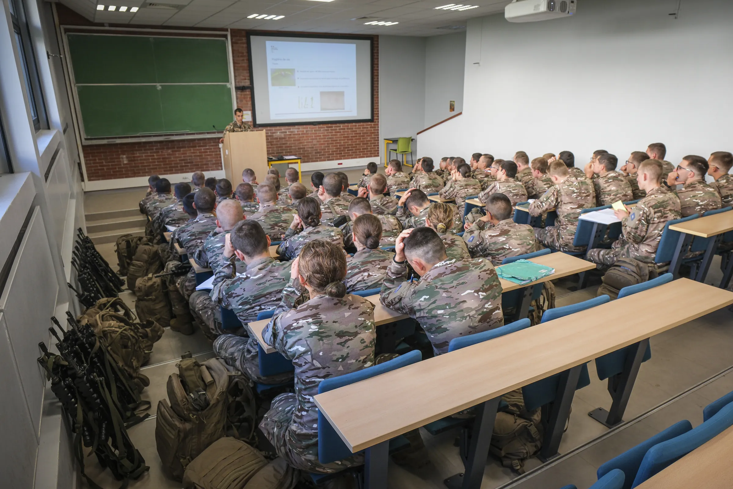A packed lecture theatre of cadets in camouflage, rifles stacked at the rear