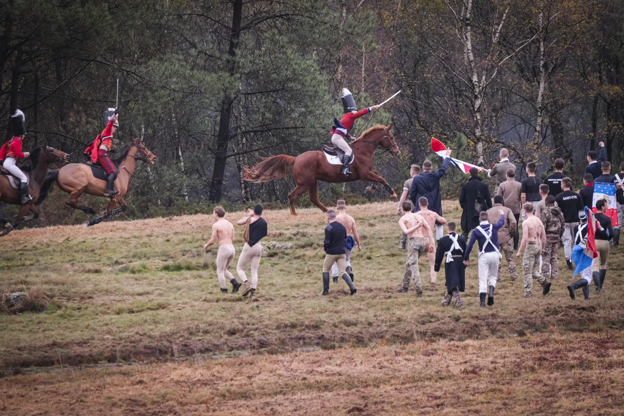 Red-coated cavalry gallops past bare-chested cadets in Saint-Cyr shirts charging uphill with flags