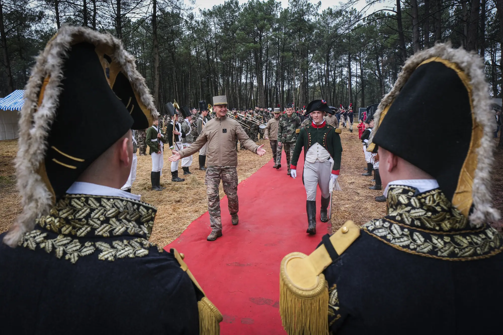 A general in camouflage is greeted on a red carpet by cadets dressed as Napoleon and his marshals