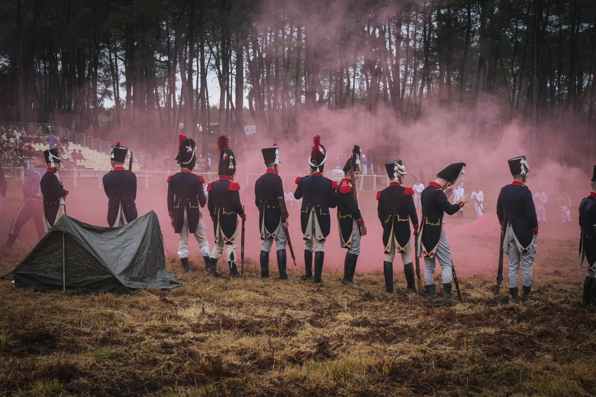 A line of reenacted grenadiers with muskets amid pink smoke before spectators