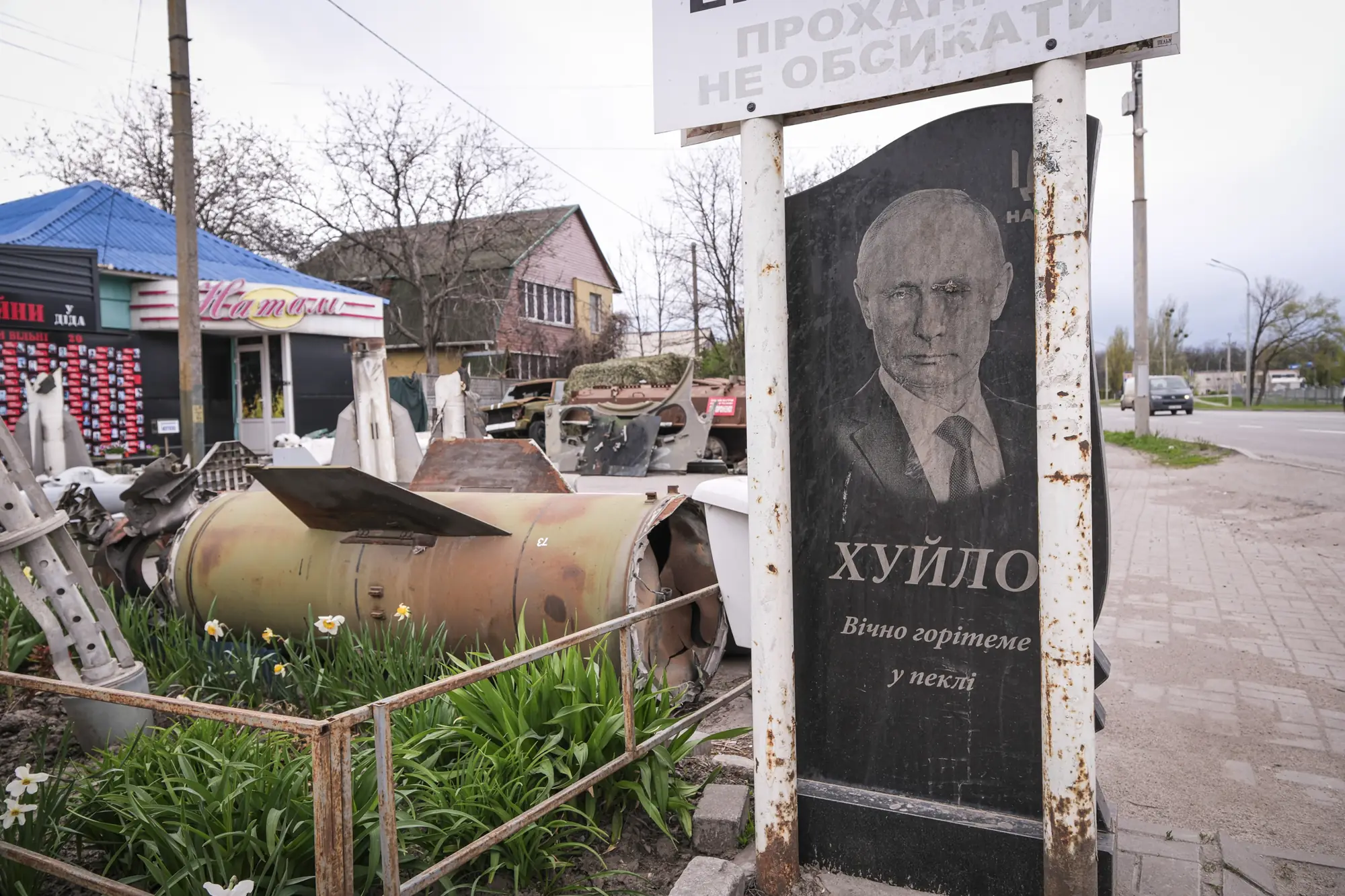 A mock gravestone of Vladimir Putin beside missile debris and destroyed armour, with daffodils