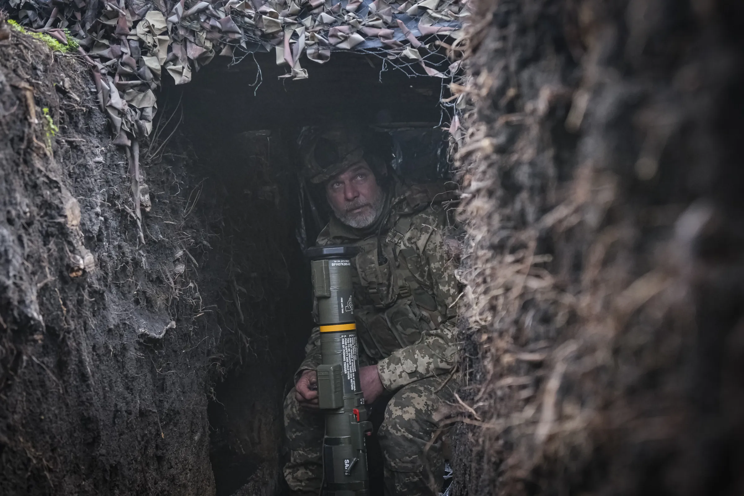 An older Ukrainian soldier crouches in a narrow camouflaged trench holding an AT4 anti-tank launcher